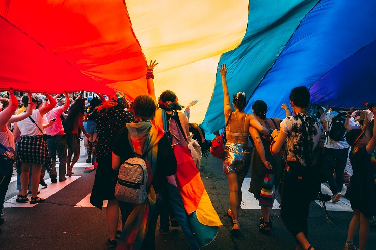 plusieurs personnes défilent sous un drapeau arc-en-ciel LGBTI+